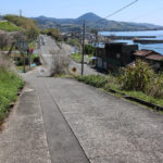 下り坂の街道が国道34号線と交差。交差後の街道が正面右側に見える。大村湾と小さな港、棚田、鉢巻山がみえる(千綿宿郷23-2付近)